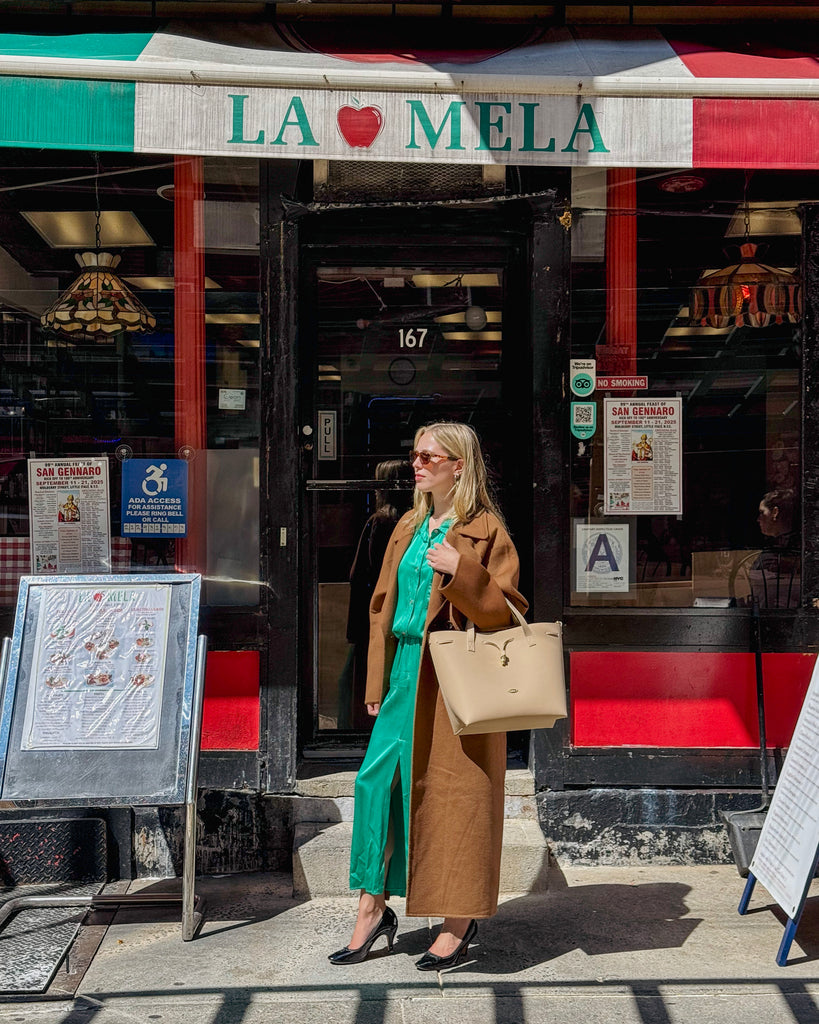 Woman in a brown coat and green dress standing outside a store with a colorful awning, holding the beige handbag